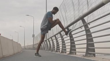Düşük açılı, uzun boylu, formda, spor giyimli Afro-Amerikan bir adam şehirde köprüde egzersiz yaparken bacak esnetme egzersizi yapıyor.