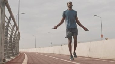 Genç atletik siyahi adamın güneşli bir günde koşu pistinde ip atlarken tam boy yavaş çekimi.