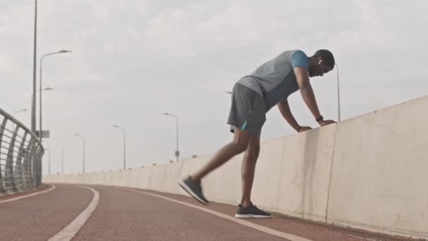 身穿运动服和无线耳机的年轻的非洲裔慢跑者在跑道上做腿部运动 在阳光灿烂的早晨在室外训练的全长慢镜头 — 图库视频影像