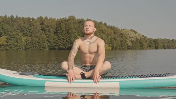 晴れた日に一人で湖でボード上の蓮のポーズで座って 目を閉じて瞑想若い入れ墨の男のスローショット — ストック動画