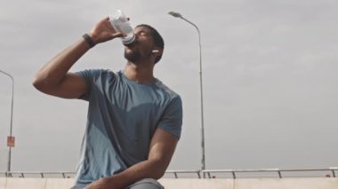 Siyah adam sabah antrenmanından sonra dinleniyor, su içiyor ve kablosuz kulaklıkla müzik dinliyor.