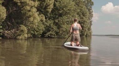 Spor mayo giymiş beyaz bir kadının göl kıyısında yemek masasında kürek çekerken yavaş yavaş arkadan çekilen görüntüsü.