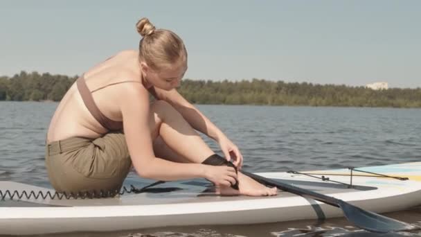 白种人年轻女子坐在湖中的冲浪板上 与人交谈 将冲浪带绑在脚踝上的慢镜头 — 图库视频影像
