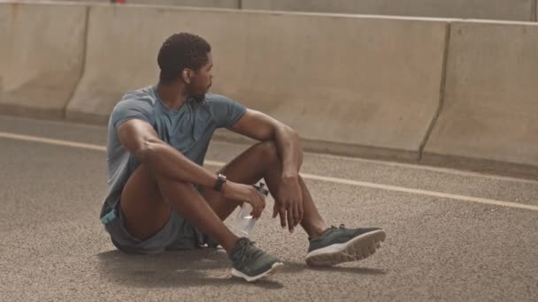 坐在城市桥饮用水边的疲惫的非裔美国人在运动后休息的慢镜头 — 图库视频影像