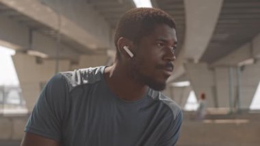 Spor giyim ve kablosuz kulaklıklar takan Afro-Amerikan sakallı adamın kameraya doğru yavaşça yakın plan çekimi.
