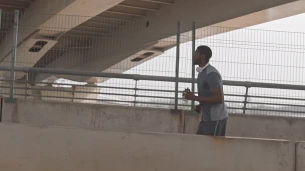 穿着运动服和耳机的黑人运动员在城市环境下从桥上慢跑时的侧视中等速度 — 图库视频影像