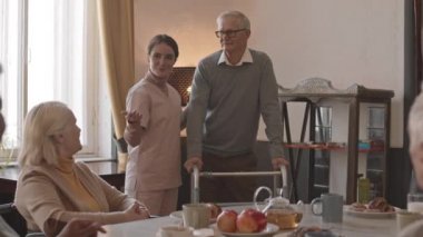 Yavaş çekilen orta sınıf bir hemşire yaşlı gri saçlı adamı huzurevindeki masada oturan yaşlı insanlarla tanıştırıyor.