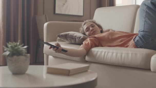 中出しの退屈若いです白人女性でキャサリーウェアスイッチングチャンネルオンテレビ寝そべっている上のソファの上にリビングルーム — ストック動画