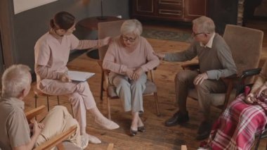 Yaşlı bir kadının diğer yaşlılar ve uzmanlarla grup terapi seansı sırasında ağlarken yavaş çekimde huzurevinde oturup ona destek olurken görüntüsü.