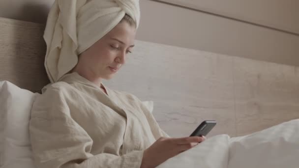 Kafasına Bornoz Havlu Geçirmiş Rahat Bir Yatakta Oturmuş Akıllı Telefondan — Stok video