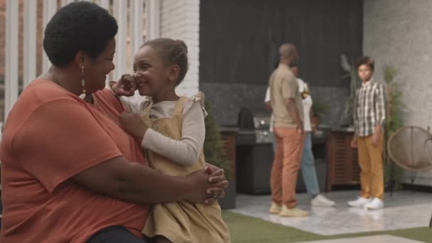 中等年龄的非洲裔美国妇女和她可爱的小孙女坐在一起 谈笑风生 身后站着模糊的家人 — 图库视频影像