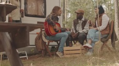 Yavaş çekimde, arkadaşlara gitar çalan bir adam yaz ormanında tekerlekli küçük bir evin yanında bira şişeleriyle birlikte oturuyor.