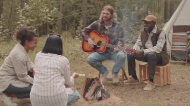 Kareli tişörtlü, beyaz bir gencin ormanda kamp ateşinde yan yana oturan çok ırklı arkadaşlarına gitar çaldığı yavaş çekim.