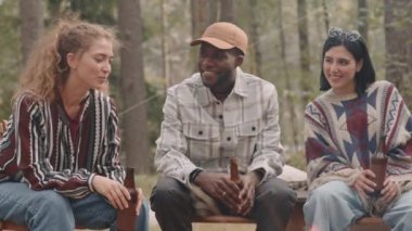 Ormanda kamp ateşinin yanında oturup sohbet ederken iki genç kadın ve Afro-Amerikalı adamın gülüp bira içtiği yavaş çekim.