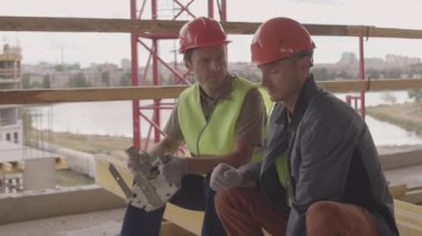 Turuncu miğferler takan iki beyaz inşaat işçisinin şehir manzarasının ön tarafındaki bir kirişe oturmuş, konuşuyor ve metalik mekanik parçalar tutuyor, gülümsüyor.