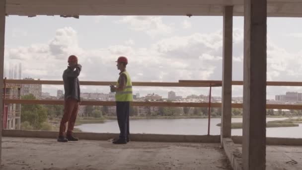 建設中の建物の内側の端に立ってヘルメットを身に着けている2人の白人労働者の広い追跡左夏の日に話して — ストック動画