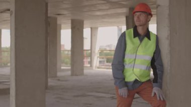 Orta yaşlı, kasketli ve yansıtıcı yelek giyen, inşaat halinde duran, beton direğe dayanan orta yaşlı beyaz bir adam.