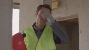 Mavi gözlü Kafkas inşaat işçisi yansıtıcı yelek giyiyor, sıkı çalıştıktan sonra çıkartıyor ve geri takıyor, beton sütun inşaatında ayakta duruyor.