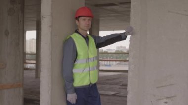 Orta yaşlı, yansıtıcı yelek giyen, inşaat halindeki binada dikilip beton sütunlara yaslanan, mesafeye bakan orta yaşlı beyaz bir adam.