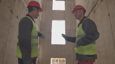 Düşük açılı orta boylu, orta yaşlı iki beyaz erkek yansıtıcı yelek ve turuncu başlık giyerek çok katlı bina çerçevesinde konuşuyor ve el kol hareketleri yapıyorlar.