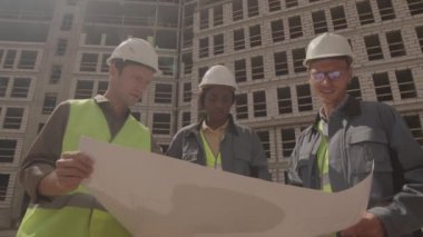 Düşük açılı üç farklı mimarın üniformalı, başlıklı, planlı kağıt tutan, inşaat halindeki çok katlı binanın ön planında tartışan