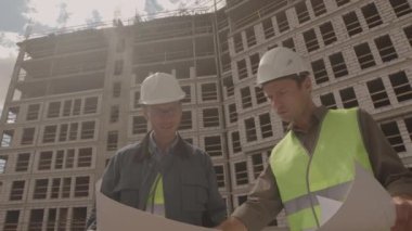 Düşük açı, iki beyaz erkek mühendisin üniforma giyip, baret takıp, elinde planlı kağıt tutarak inşaat halindeki çok katlı binanın ön planında tartışmalarıydı.