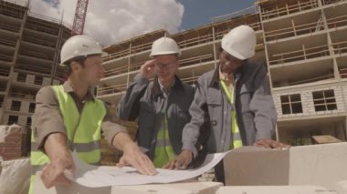 Üç çok ırklı mimarın iş kıyafetleri giyip, başlık takıp, planlı kağıda bakıp, inşaat halindeki çok katlı binanın ön planında konuşmalarının orta derecede düşük açılı görüntüsü.