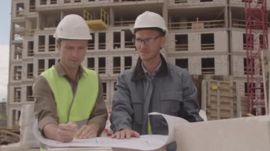 Kafkasyalı iki mimarın iş kıyafetleri ve sıkı şapkalar giymesi, planlı kağıda bakması, yapım aşamasındaki çok katlı binanın ön planında tartışması.