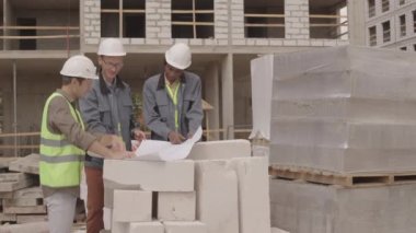 Çok ırklı üç mimarın iş kıyafetleri ve sıkı şapkalar giyip, planlı kağıda bakıp, yapım aşamasındaki çok katlı binanın ön planında tartışmalarının orta uzunluğunda.