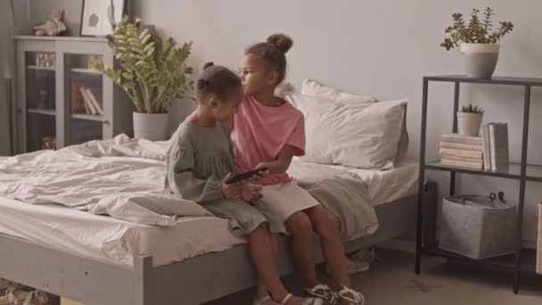 2人のかわいい5と8歳のアフリカ系アメリカ人の女の子の遅いショットは ベッドの上に座っている居心地の良いアパートでスマートフォンでゲームをプレイし チャット — ストック動画