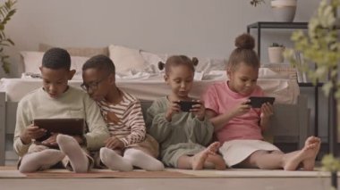 Dört Afro-Amerikan okul öğrencisi kız ve oğlanın rahat yatak odasında oturup aletlerle sanal oyunlar oynamalarının yavaş çekimleri.