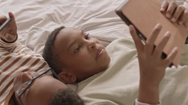 从上往下拍的中特写镜头 慢慢地 一个可爱的非洲裔美国少年双胞胎男孩把头靠在一起躺在床上玩小游戏和聊天 — 图库视频影像