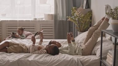 Yavaş çekimde Afro-Amerikan ikiz çocuklar akıllı telefon ve dijital tabletle oyun oynuyorlar rahat evlerinde yatakta uzanıyorlar ve 5 yaşındaki kız kardeşleri arka planda oturan akıllı telefonunu kullanıyor.