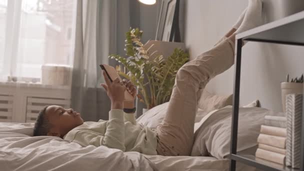 用数码平板电脑拍摄穿着便服 双腿高高地躺在床上的非裔美国少年的侧视慢镜头 — 图库视频影像