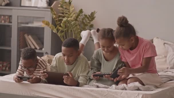 ゆっくりショットの4かわいいアフリカ系アメリカ人の男の子と女の子のゲームで電子ガジェット座っていると横たわっでベッドで居心地の良いアパート — ストック動画