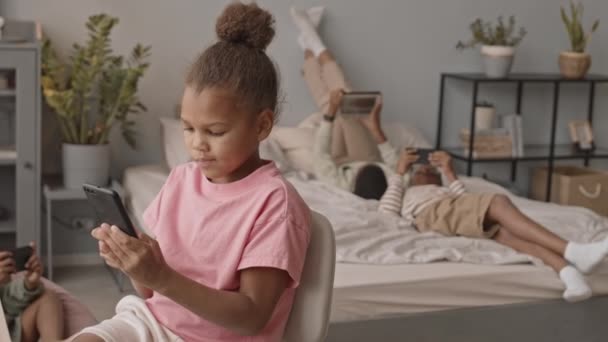 中慢镜头拍摄的8岁的非洲裔美国女孩在家里打滚或玩智能手机 而她的朋友或兄弟姐妹也在背后使用小工具 — 图库视频影像