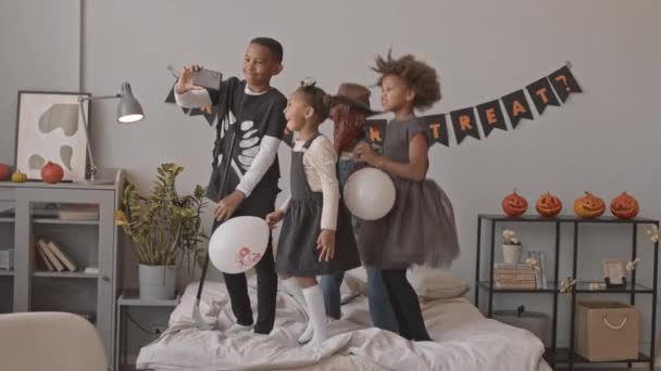 ハロウィンのホームパーティーで一緒に楽しい衣装で楽しいアフリカ系アメリカ人の子供たちのスローショット ベッドでジャンプし スマートフォンで自分自身のビデオを撮る — ストック動画