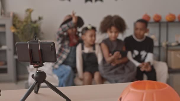 ハロウィンパーティー中にベッドの上に座っているスマートフォンでビデオチャットを持つハロウィーンの衣装で4陽気で遊び心のあるアフリカ系アメリカ人の子供たちのスローショット — ストック動画