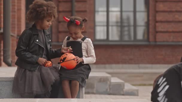 两个穿着万圣节服装的可爱小女孩坐在户外用南瓜桶装糖聊天的慢镜头 — 图库视频影像