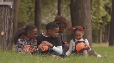 Dört neşeli Afro-Amerikalı çocuğun cadılar bayramı kostümlü dışarıda çimenlerde oturup çene çalarken ve kahkahalar atarken yavaş çekimleri.