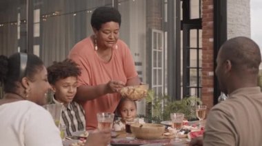 Orta uzunlukta mutlu bir Afrikalı büyükanne, modern evin arka bahçesinde oturan aile üyelerine salata servisi yapıyor.