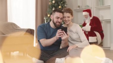 Oturma odasında oturan, akıllı telefon kullanan ve gülümseyen genç, beyaz heteroseksüel çift Noel Baba hediyelerini arka plandaki Noel ağacının altına yerleştiriyor.