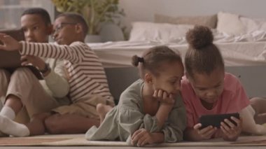 Akıllı telefon ve dijital tabletle oyun oynayan Afro-Amerikan öğrencilerin rahat bir dairede birlikte otururken yavaş çekimleri.