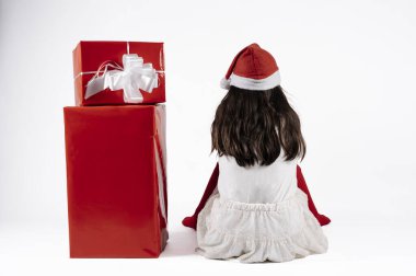 Beyaz arka planda kırmızı hediyelerin yanında sırt üstü oturan Noel Baba şapkalı küçük kız. Noel konsepti