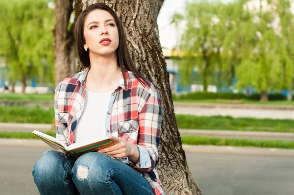 Mujer pensativa sentada bajo el árbol y sosteniendo un libro — Foto de Stock