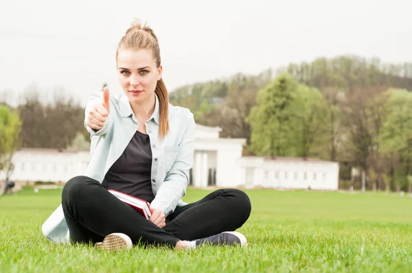 Young woman with book showing thumb up and sitting outdoor — Stock fotografie