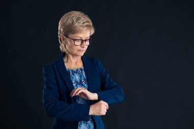 Yaşlı sekreter mavi ceketini koyu bir arka planda morumsu gözlük takıyor.