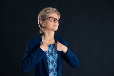 Yaşlı sekreter mavi ceketini koyu bir arka planda morumsu gözlük takıyor.