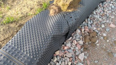 Bir inşaat sahasında toprak ve çakıl ile çevrili bodrum su geçirmez ya da drenaj için kullanılan siyah gamzeli zarın yakın çekimi..