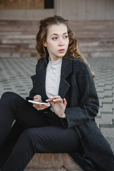 卷曲头发的女孩穿着黑色外套坐在街上 看着电话 — 图库照片